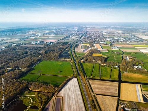 Aerial panoramic view of the Dutch countryside featuring farmland, green fields, forests, and a highway stretching through the landscape toward a distant town.