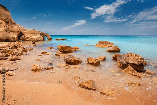 Fototapeta Naklejka Na Ścianę i Meble -  Crystal clear turquoise water gently washing golden sand and rocks on a mediterranean beach with cliffs and arches on a sunny summer day