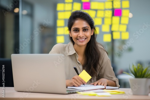 Confident young businesswoman writing on sticky notes at modern office desk with laptop and planner, planning company strategy