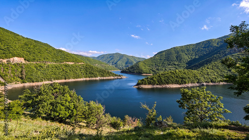 Panoramic view of Vacha Reservoir in Bulgaria, surrounded by lush green mountains and calm blue water under a bright sky.