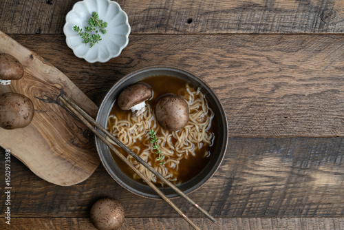 Hot noodle soup with mushrooms and thyme on a wooden table with cutting board and chopsticks
