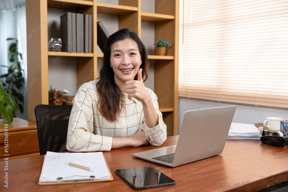 Asian young adult woman sitting at wooden desk in home office smiling confidently and showing thumbs up gesture, wearing checkered shirt beside laptop, clipboard, tablet and coffee mug Asian young adult woman sitting at wooden desk in home office smiling confidently and showing thumbs up gesture, wearing checkered shirt beside laptop, clipboard, tablet and coffee mug