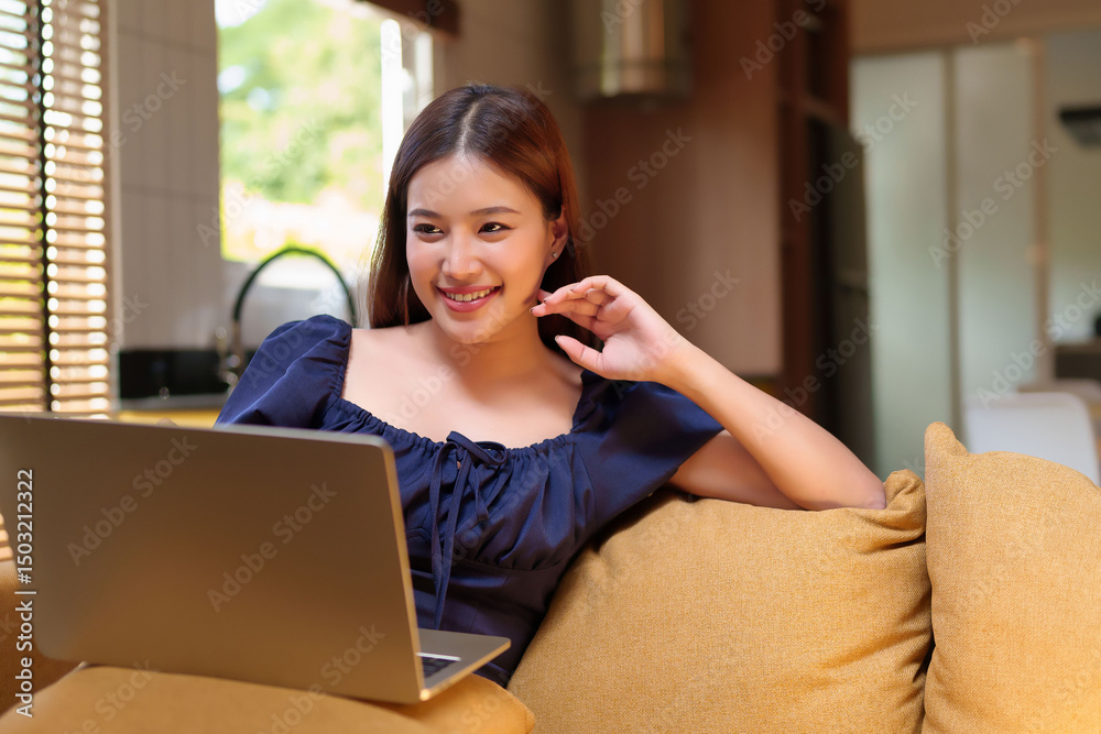 Naklejka premium Asian woman using laptop on couch Adult smiling and working from home in a cozy living room Happy face working on her computer