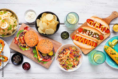 Summer food table scene with hot dogs, hamburger and sides. Above view over a white wood background.