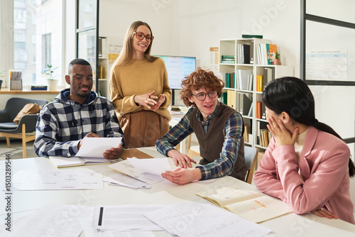 Students engaging in group study session reviewing foreign languages with materials and textbooks around large table. Instructor standing nearby, assisting and smiling