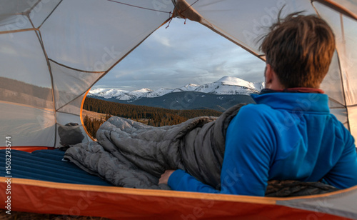 View from inside a tent of a man lying in a sleeping bag, looking at snow-covered mountain peaks at sunset.