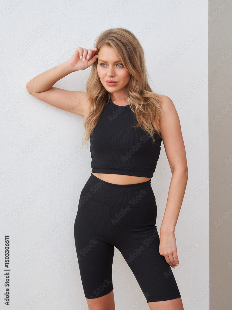 Naklejka premium Young woman in a black athletic outfit poses confidently, hand touching her hair. Her top is sleeveless and paired with bike shorts. Her expression is calm and focused. Casual and stylish