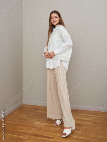Smiling woman with long brown hair in a white oversized shirt, beige sweater vest, and pleated wide-leg pants stands on a hardwood floor wearing white heeled sandals