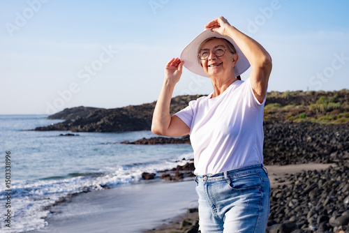 Smiling carefree senior woman in white summer hat looking at new day from the sea beach enjoying freedom, vacation or retirement concept