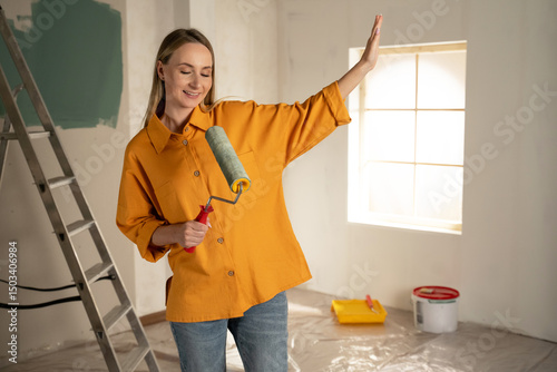 Happy house painter having fun singing and dancing while using a paint roller to redecorate a room, enjoying her work and expressing her creativity