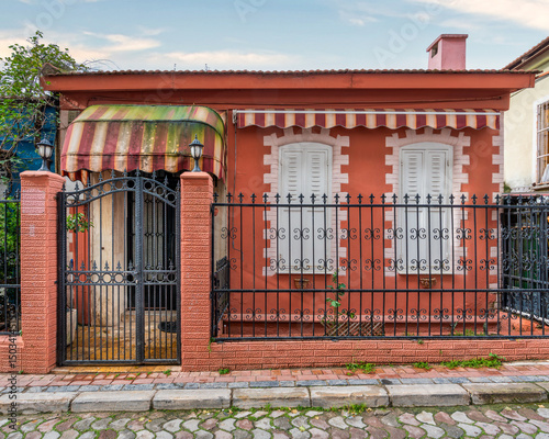 Fototapeta Naklejka Na Ścianę i Meble -  Exterior view of a small orange house with white shutters, black metal fence, and striped awnings on a cobblestone street, at daytime