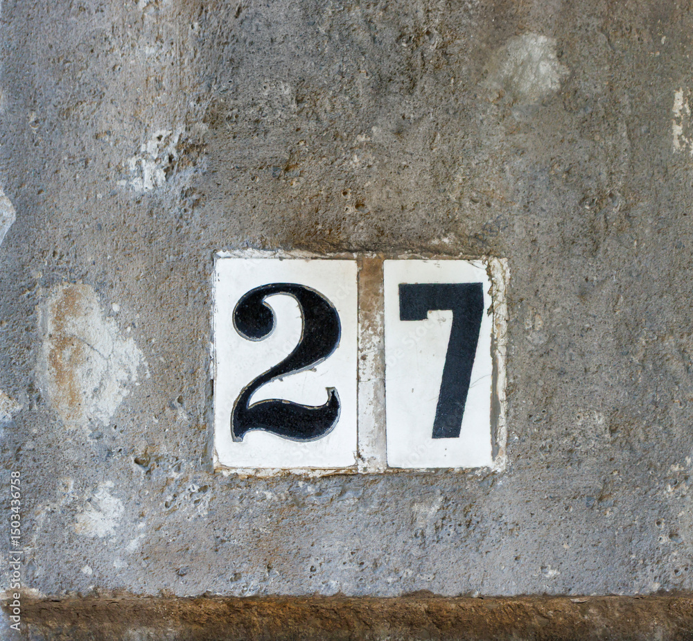 Street address number, number 27, in black on white tile inset into a dark rough stone wall. Street address number, number 27, in black on white tile inset into a dark rough stone wall.