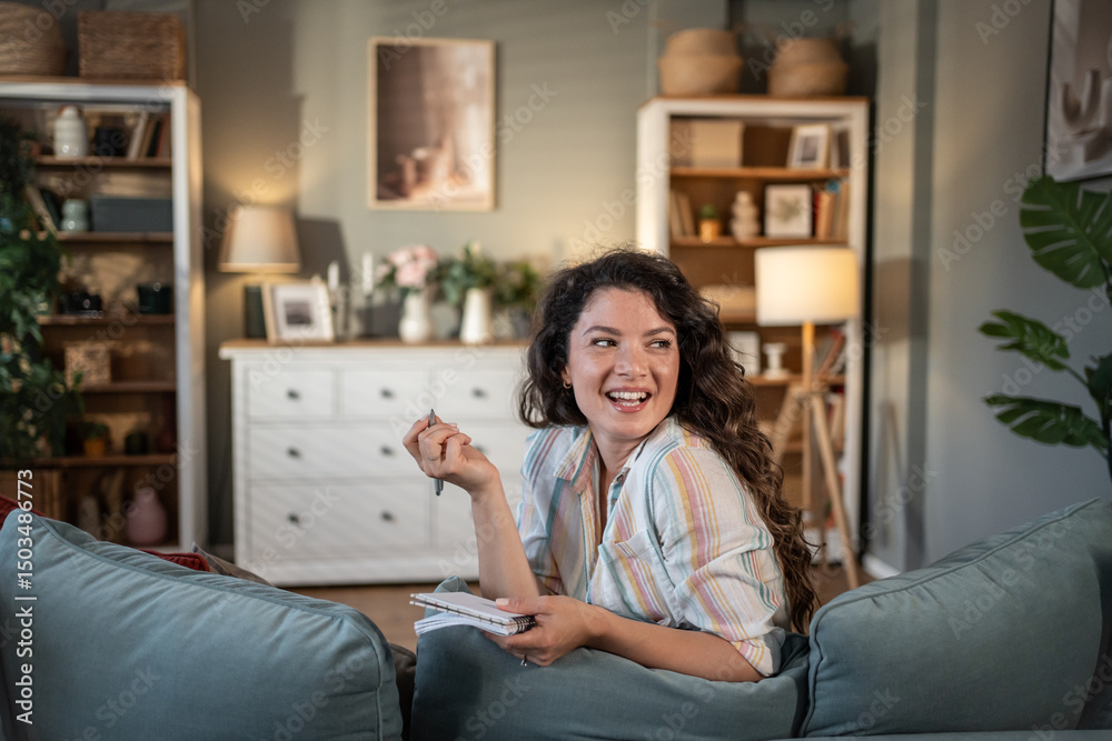 Obraz premium Young woman taking notes and smiling on sofa at home