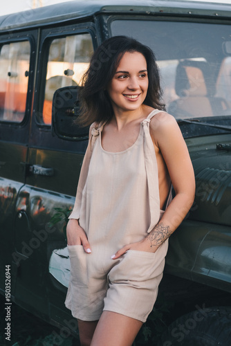 A beautiful woman in a light jumpsuit with a hat stands near a car in the village. Tinting
