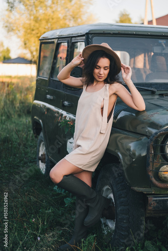 A beautiful woman in a light jumpsuit with a hat stands near a car in the village. Tinting
