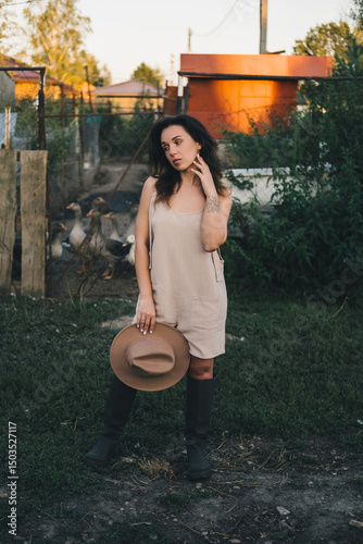 A beautiful woman in a light jumpsuit with a hat stands near a paddock with geese village. tinting