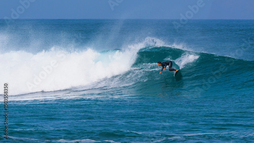 Surfer in black wetsuit making cutback turn on a curling reef break wave as offshore winds creates a dramatic arc of mist. Powerful ocean swell forms perfect waves at surf spot on island of Lanzarote.