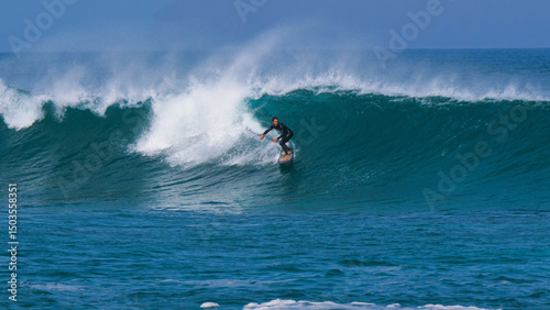 Regular footed rider surfing along a steep blue wave as offshore wind creates a dramatic spray. Powerful ocean swell forms perfect waves at surf spot on island of Lanzarote in the Canary Islands.