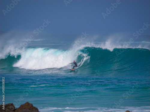 Experienced male surfer leans into a bottom turn while surfing big, clean wave. Powerful ocean swell forms perfect waves as offshore winds creates dramatic arcs of mist at surf spot on Canary Islands.