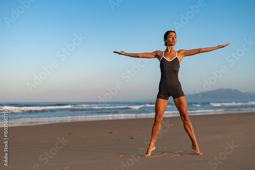 Focused athletic woman standing barefoot on the beach in a strong warrior pose at sunrise, arms stretched out wide, expressing balance, power, and connection with nature.