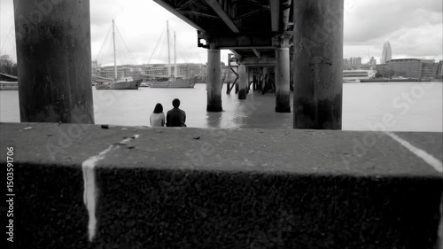 Couple sitting by the river under a bridge, boats and city buildings in the background, black and white mood, monochrome scenery