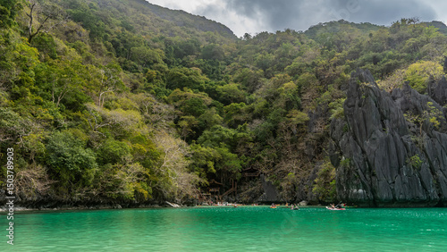 Tourist canoes float in the emerald lagoon. Picturesque coastal cliffs. Lush green tropical vegetation on the mountain slopes. Blue sky, clouds. Philippines. Palawan. Small Lagoon. Bacuit archipelago.