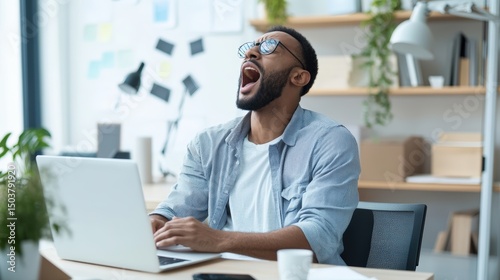 Tired Man Yawning While Working on Laptop in Modern Office Space