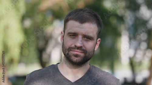 A man with a beard stands in a park setting, surrounded by green trees and natural light, looking relaxed and approachable while wearing a simple gray shirt.