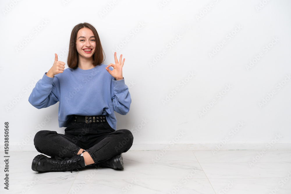 Young Ukrainian woman sitting on the floor isolated on white background showing ok sign and thumb up gesture Young Ukrainian woman sitting on the floor isolated on white background showing ok sign and thumb up gesture
