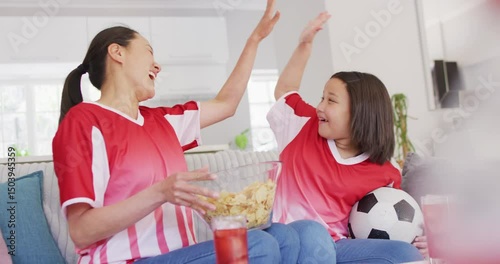 Video of happy asian mother and daughter eating chips and watching match in tv