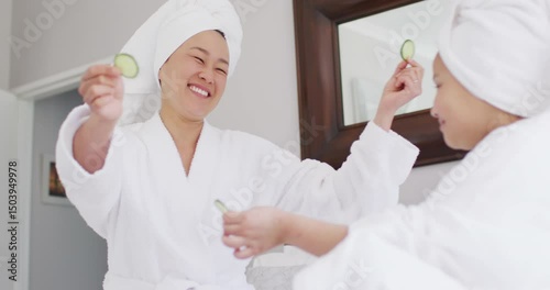 Video of happy asian mother and daughter in robes doing cucumber mask and having fun