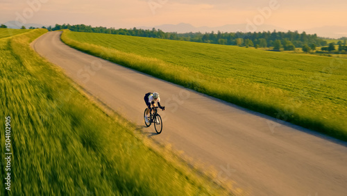 A person rides a bicycle down a winding road, surrounded by lush green fields. The camera captures a dynamic aerial view, emphasizing the sense of speed and freedom in this serene countryside setting.