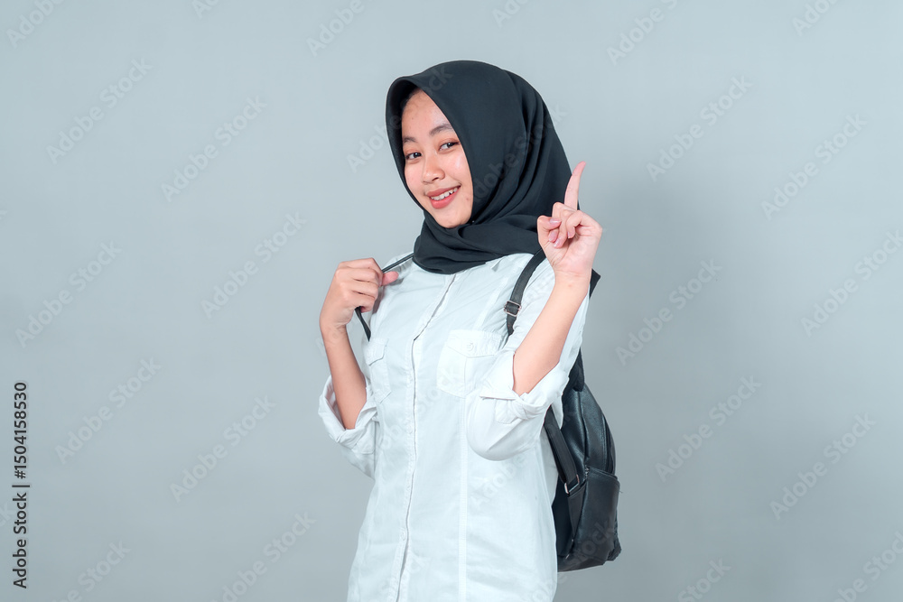 Confident young Asian woman wearing a black hijab and white shirt, holding her backpack strap and raising one finger, smiling while posing against a plain grey background. Confident young Asian woman wearing a black hijab and white shirt, holding her backpack strap and raising one finger, smiling while posing against a plain grey background.