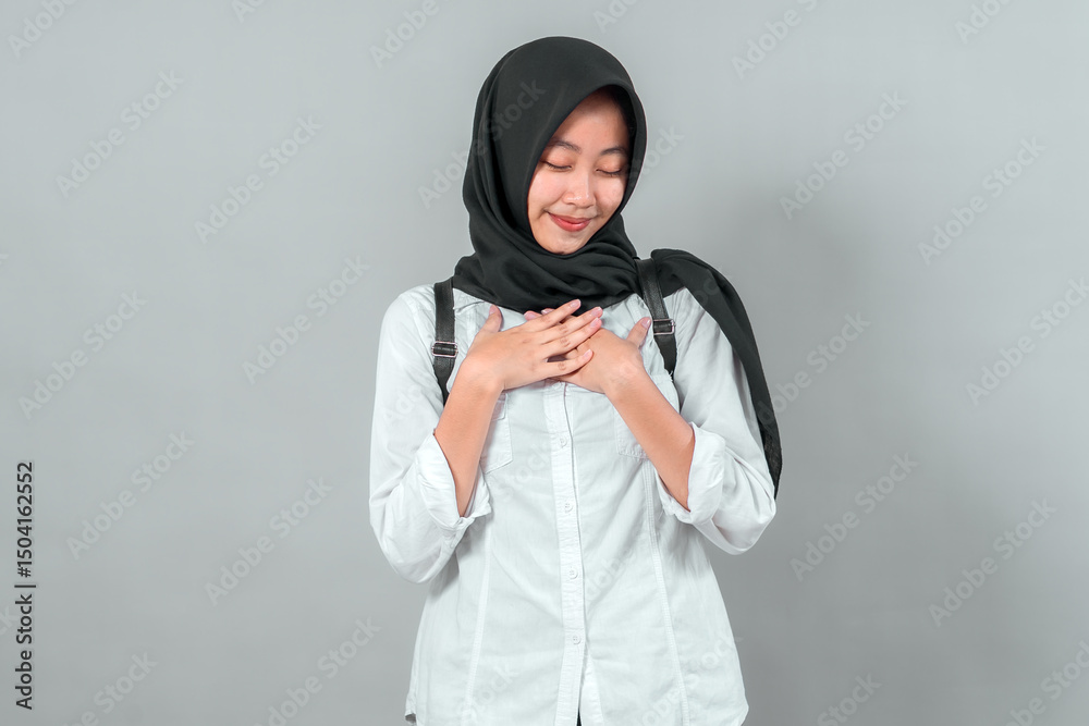 Obraz premium Modest Muslim student wearing a hijab and backpack, smiling gratefully with hands on chest. Back to school concept, expressing humility and enthusiasm in a studio shot.