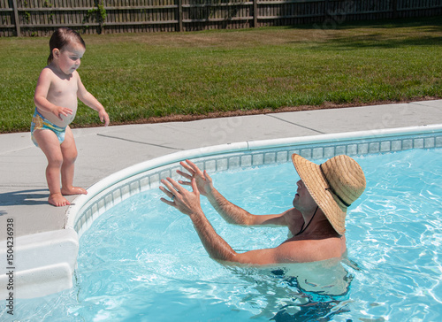 Young father coaxing his young daughter to jump into his arms in a swimming pool.  