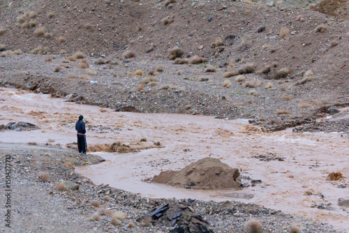 A person stands beside a muddy stream in a rugged, barren landscape near Taft, Yazd, Iran, observing the flowing water against the backdrop of rocky terrain and sparse vegetation. photo