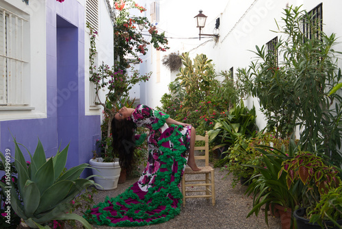 Fotografie Young and beautiful woman dancing flamenco rests one foot on an old wooden chair in a typical street of andalusia with flowers and throws her head back