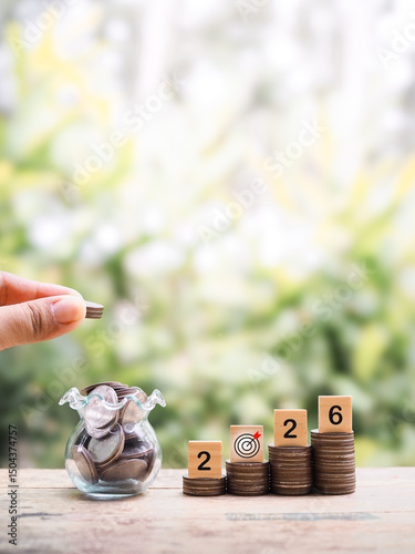 Hand putting coin into glass bottle, Wooden blocks with the number 2026 on stack of coins. The concept of saving money, Financial, Investment and Business growing in year 2026.