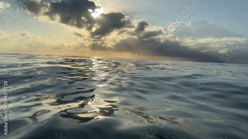 A golden sun rising over the ocean at dawn with rays beaming out from puffy clouds colouring the ocean with beautiful silvery tones. Captured in slow motion within the ocean on sunrise. 