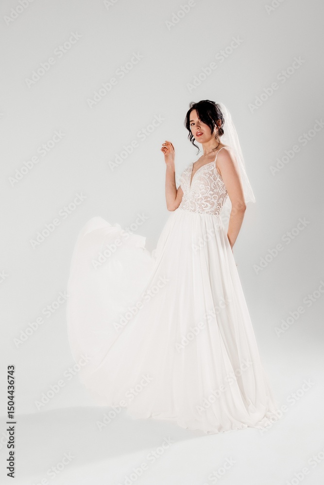 Fototapeta premium Bride in elegant white dress with flowing fabric poses gracefully in studio setting during daytime