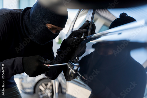 Car thief breaking into vehicle with screwdriver while wearing black mask and gloves during daylight robbery