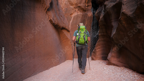 Adult man hiker with backpack and trekking poles hiking on bottom dry sandy cave riverbed in deep slot canyon with curved and smooth sandstone rocks walls of orange red color, slow motion, view back