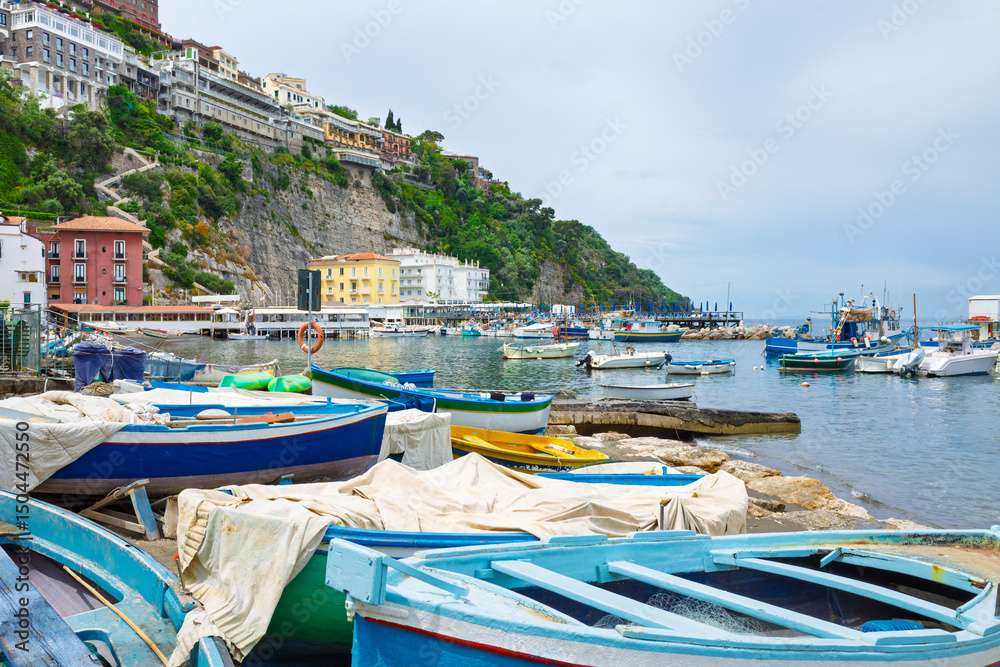 Obraz premium Colorful wooden fishing boats on shore and moored in Marina Grande harbor, Sorrento, Campania, Italy. Coastal buildings on cliffs and calm Tyrrhenian Sea under overcast sky.