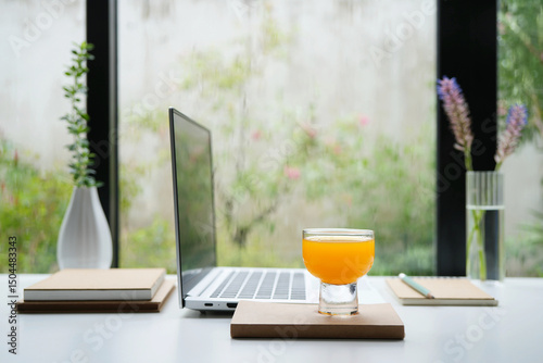 Side view of a laptop with orange juice cup and notebooks, working by the window