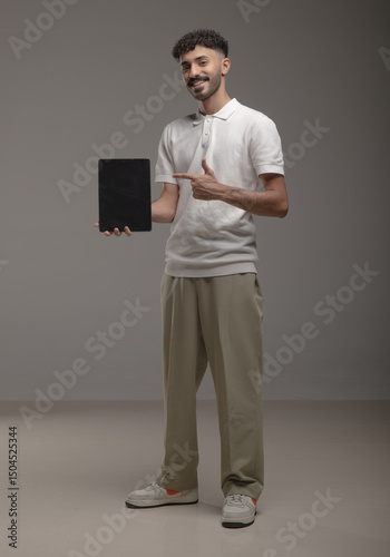 A young Saudi man in modern casual attire holding a blank white foam board for advertisement or mockup purposes, standing in a studio with a neutral background. Ideal for marketing use.