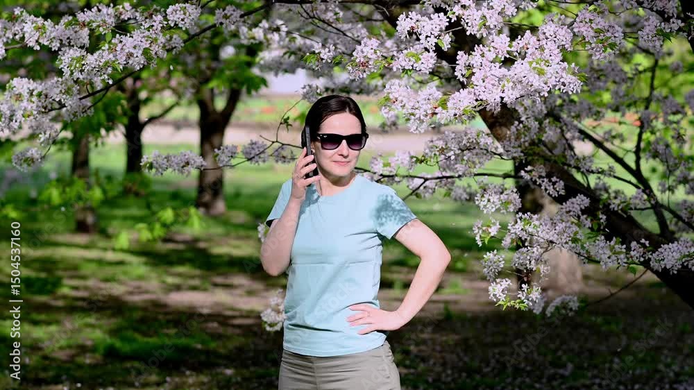 Young woman in sunglasses on video call via smartphone in blooming spring park