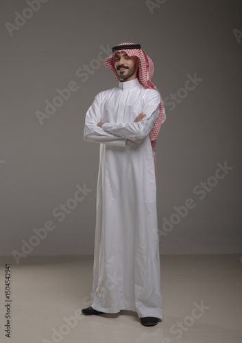 Confident Saudi man in traditional attire standing with arms crossed, expressing pride and professionalism. Studio shot with grey background, ideal for cultural and business themes.