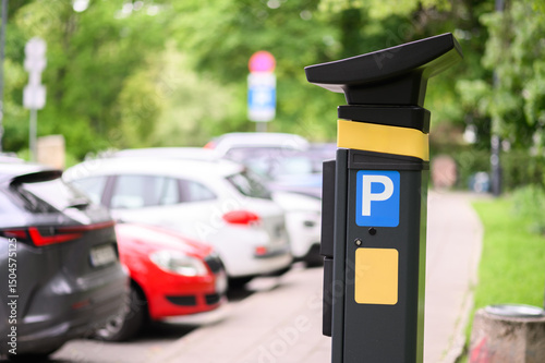 Fototapeta Naklejka Na Ścianę i Meble -  A parking meter is prominently positioned next to several vehicles in an urban parking area, surrounded by green trees and sunlight