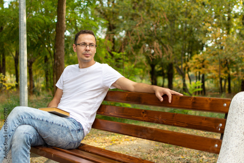 A young guy in glasses is sitting on a bench in the park with a book in his hands