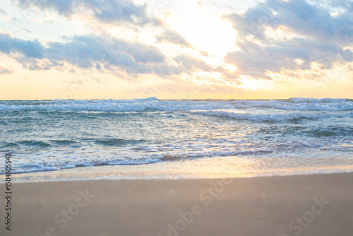 Golden sunlight shining through clouds over calm ocean with soft waves lapping at sandy beach